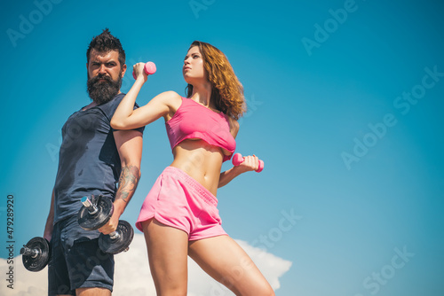 Sporty couple workout with dumbbells. Strong man and sexy woman working out with dumbbells on sky background. Muscular man and woman training arms workout using heavy dumbbells.