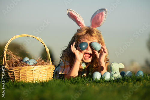 Cute excited bunny child boy with rabbit ears. Child with easter eggs in basket outdoor. Boy laying on grass in park. Easter egg hunt. Fynny kids portrait. photo