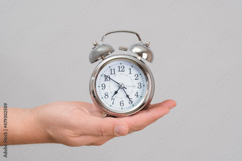 A man's hand holds a chrome alarm clock at arm's length, gray background A man's hand holds a chrome alarm clock at arm's length, gray background