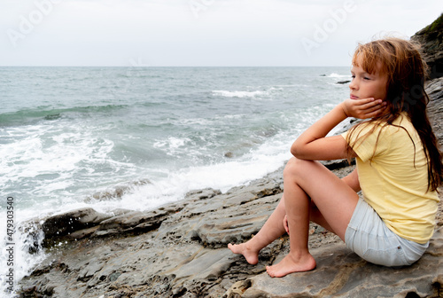 Child girl sits on the rocks by the ocean. White waves. Adventure by the sea.