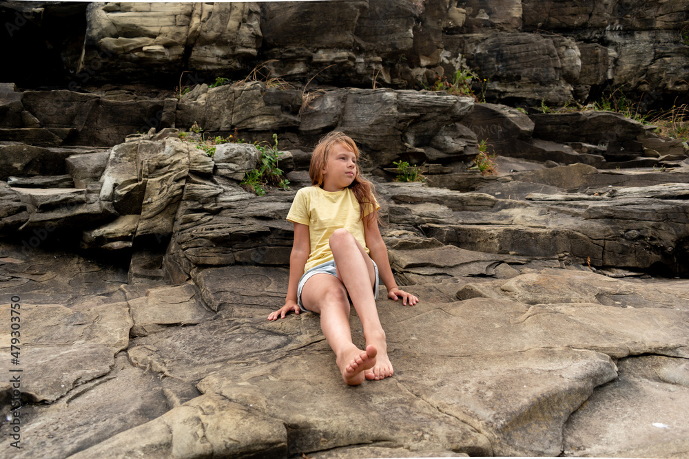 Child girl sitting on rocks by the ocean. Adventure by the sea. Wind in your hair. Child girl sitting on rocks by the ocean. Adventure by the sea. Wind in your hair.