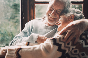 Old senior couple in love hug and embrace with romance together at home with outside view in windows background - happy mature retired people lifestyle enjoying caring each other and smiling