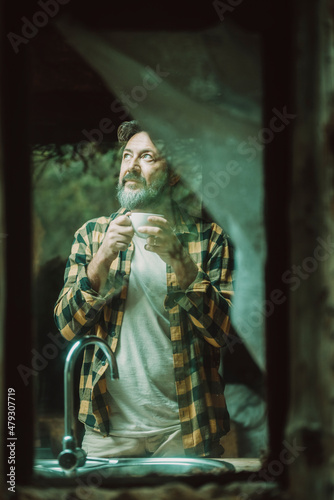 Mature adult man at home viewed from outside thrugh the glass winws drinking coffee and looking around - alternative rural lifestyle people hipster clothes and beard photo