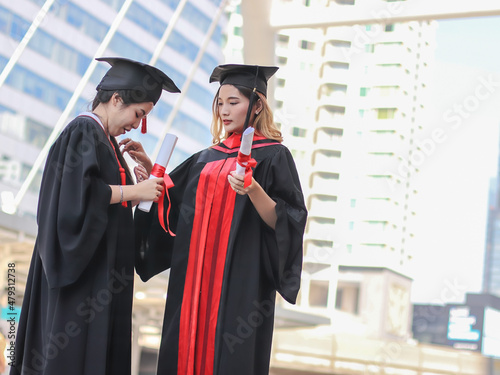  Two cheerful graduated women in graduation gowns, helping each other dress up, smiling happily. Education, successful and friendship concept.