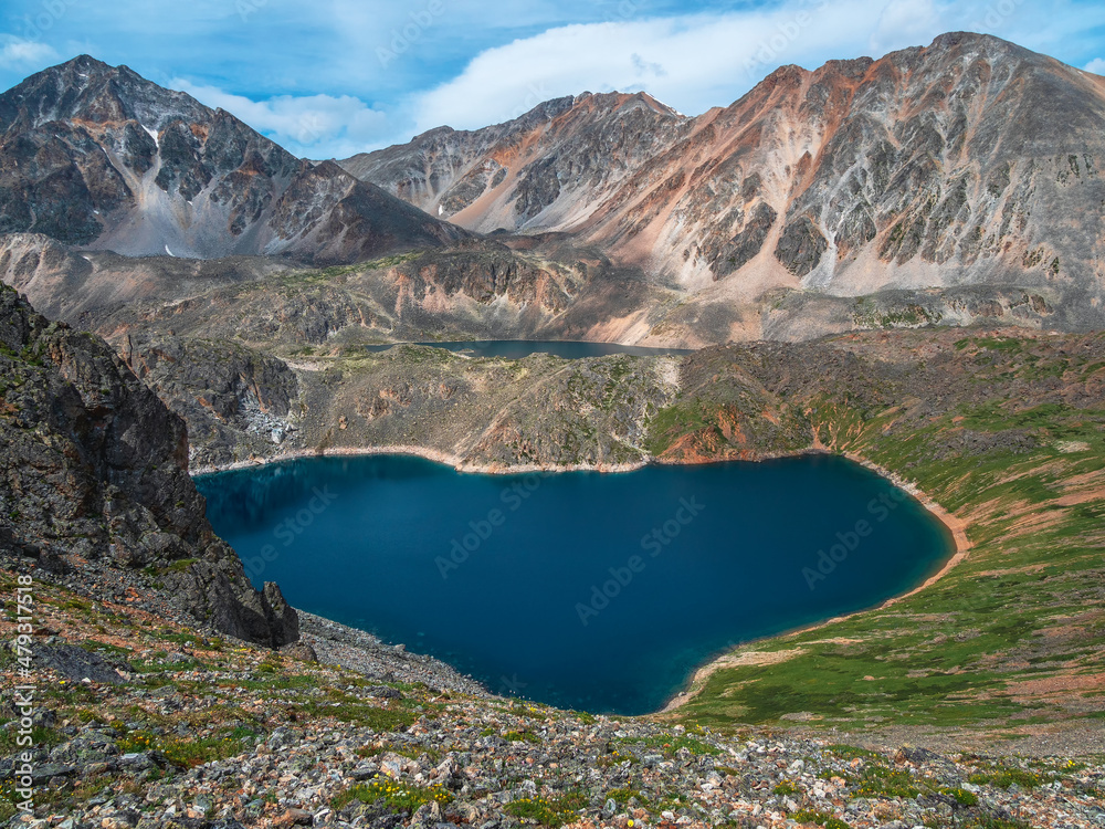Amazing view to beautiful blue mountain lake in bright sun. Heart shape blue alpine lake among sunlit green hills and large mountains under sun in cloudy sky.