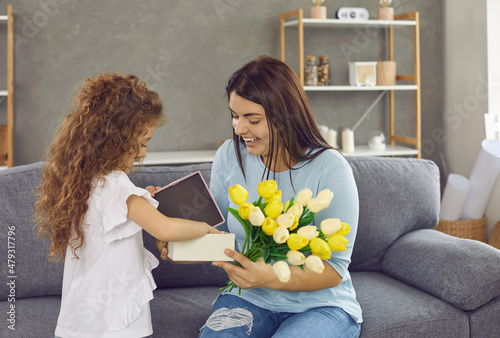 Lifestyle moment. Little daughter congratulates her loving mother on holiday by giving gifts and flowers. Happy young excited woman who is delighted with gift looks at him sitting at home on couch.