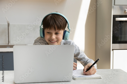 Focused smiling teen school student with headphones and dental braces watching studying course, virtual lesson, webinar, teachers class on laptop, doing homework in home kitchen, writing notes