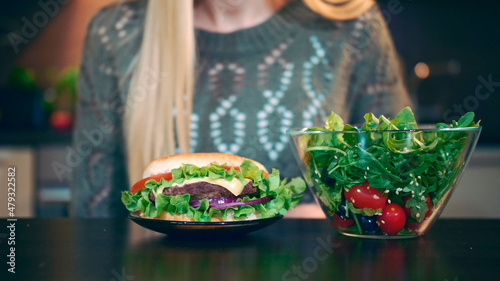 Crop view of Young lady preferring hamburger to salad. Attractive young woman choosing to eat healthy hamburger for breakfast while sitting at table in stylish kitchen.
