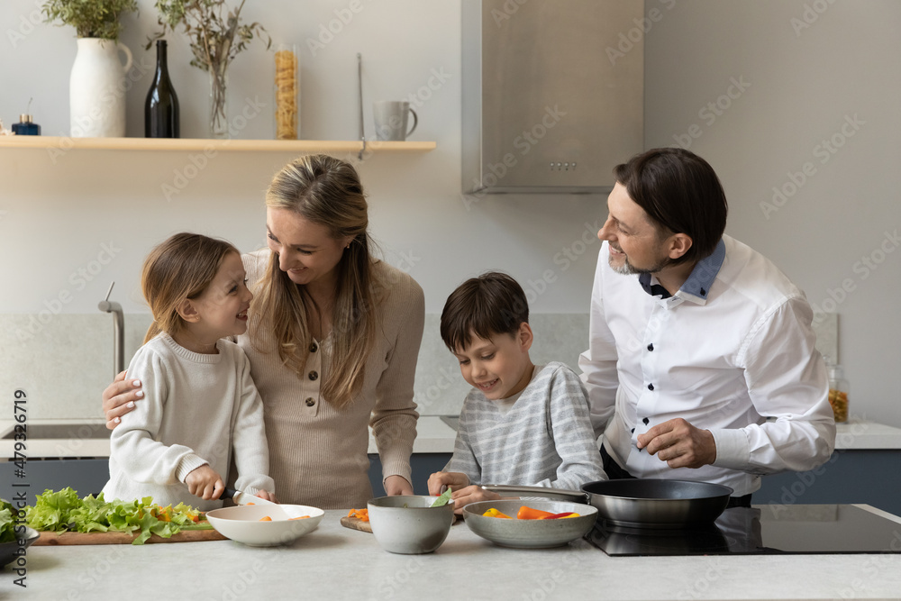 Happy parents and two sibling kids preparing lunch, dinner together, cooking healthy meal from natural ingredient, slicing salad, heating frying pan, talking, laughing, having, fun