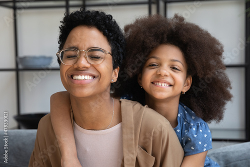 Happy African mom and cute curly haired daughter kid hugging at home, laving fun, looking at camera, smiling. Joyful mother piggybacking girl on couch, laughing, enjoying motherhood, family leisure © fizkes
