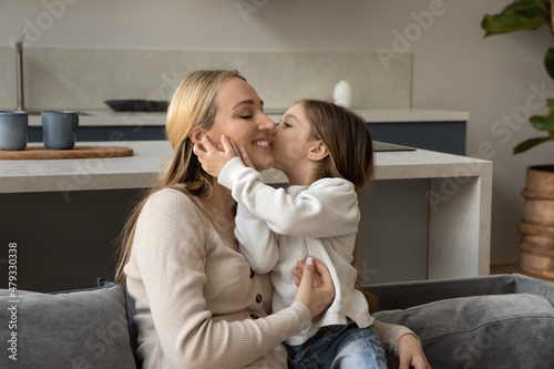 Adorable loving girl kissing happy smiling mom with love, gratitude. Mother cuddling, holding cute kid in arms, resting on couch, enjoying time with child at home. Family, motherhood concept
