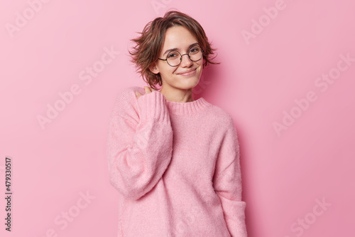 Foto Photo of satisfied European woman with bob hairstyle smiles gently keeps hand on neck glad to hear good news wears soft jumper round spectacles isolated over pink background