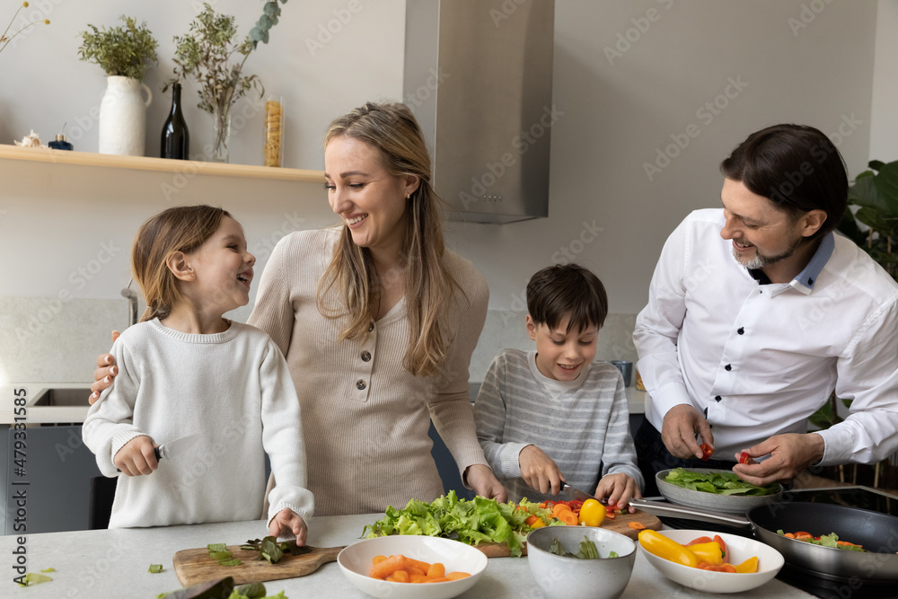 Joyful cute sibling kids helping parents to prepare dinner in home kitchen, cutting fresh vegetable at table, laughing, talking, chatting. Happy family couple teaching children to cook Joyful cute sibling kids helping parents to prepare dinner in home kitchen, cutting fresh vegetable at table, laughing, talking, chatting. Happy family couple teaching children to cook