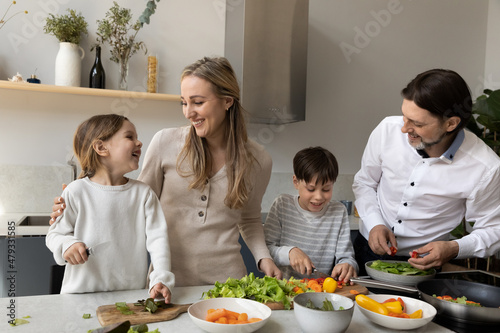 Joyful cute sibling kids helping parents to prepare dinner in home kitchen, cutting fresh vegetable at table, laughing, talking, chatting. Happy family couple teaching children to cook