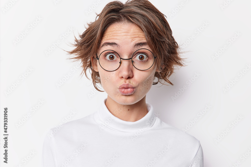 Portrait of surprised dark haired woman pouts lips as if wanting to kiss someone wears round spectacles for vision correction casual jumper isolated over white background. Human face expressions