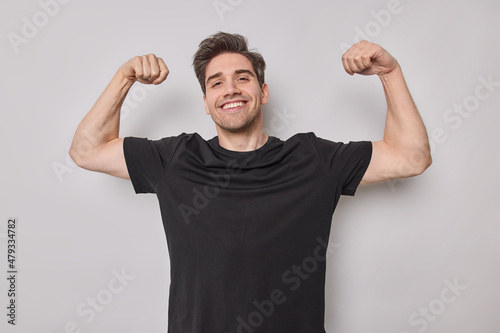 Sportive cheerful dark haired man shows biceps muscles raises arms demonstrates results of training in gym wears casual black t shirt poses against white background being strong and powerful.