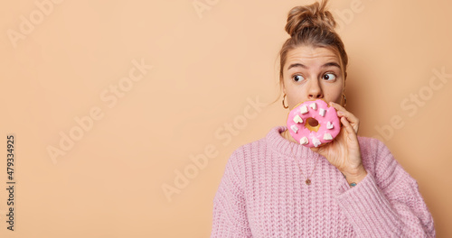 Indoor shot of surprised woman with hair gathered in bun covers mouth with delicious doughnut wears knitted jumper isolated over beige background blank empty space for your promotion or advert