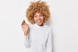 Happy woman with curly bushy hair keeps hand raise smiles sincerely expresss positive emotions wears casual turtleneck isolated over white background hears something funny. Happiness concept