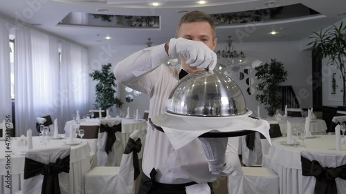 A cheerful male waiter is having fun in a restaurant, he puts a metal lid on his head while standing in a restaurant.