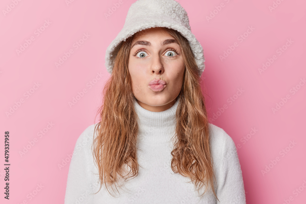 Portrait of impressed lovely young woman keeps lips folded blows kiss at camera has attractive look long hair wears fur panama and turtleneck isolated over pink bckground. Human face expressions