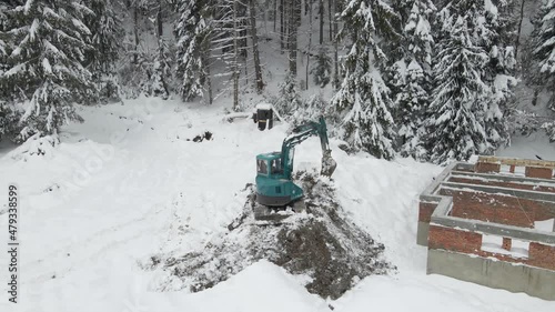 Single-bucket excavator working at a construction site in the picturesque location near the mountain forest in winter. Leveling the ground for the future building with an excavator in the snow. Dredge