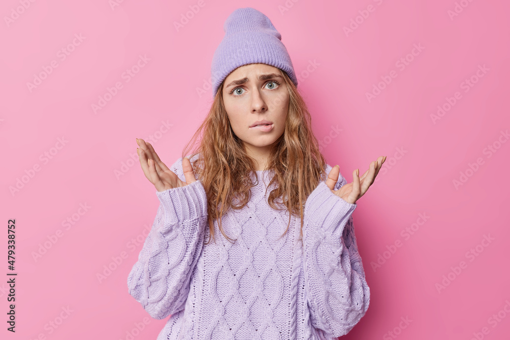 Puzzled indignant European woman with wavy hair spreads hands bites lips doesnt know what to do has clueless expression wears knitted sweater and hat poses against pink background. I dont care Puzzled indignant European woman with wavy hair spreads hands bites lips doesnt know what to do has clueless expression wears knitted sweater and hat poses against pink background. I dont care