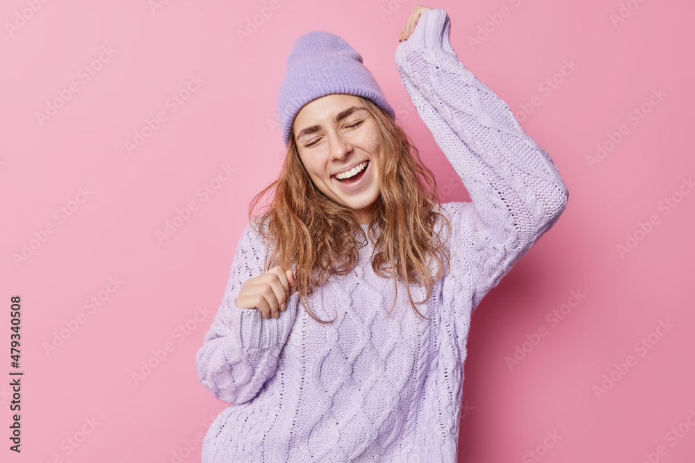 Happy beautiful party girl moves with rhythm of music dances against pink background shakes arms and sings song wears hat and sweater isolated over pink background feels like champion has fun
