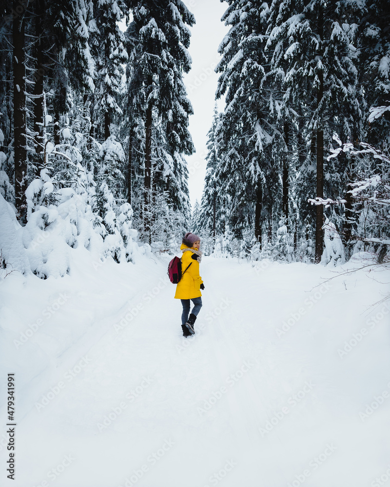 Young female in eye-catching yellow winter jacket walking trough forest full of snow. Girl in winter wonderland. 