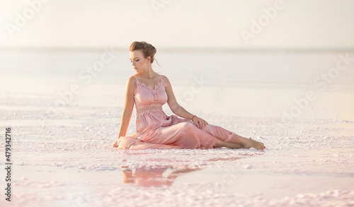 a beautiful young girl is sitting in a pink dress on the shore of a salt pink lake