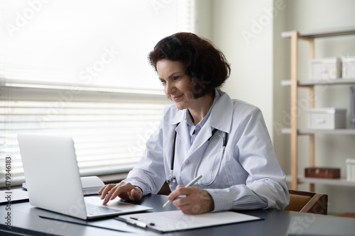Happy old mature 60s female doctor general practitioner doctor physician working on computer, handwriting notes in disease history on clipboard paper, writing healthcare prescription in clinic office.
