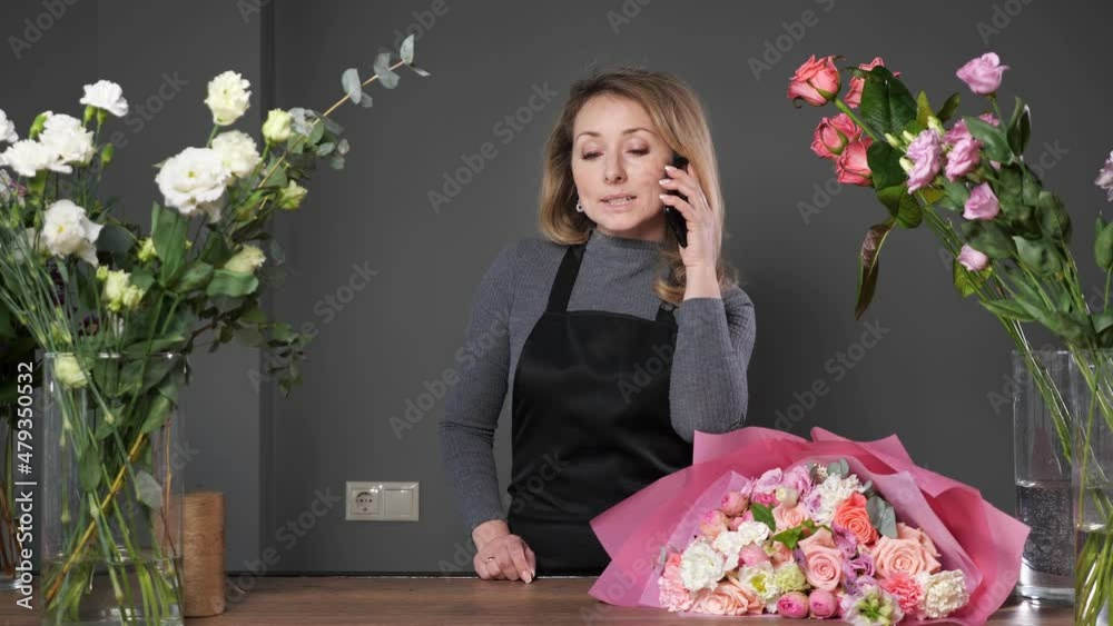 Florist woman in black apron informs customer via mobile phone about arranged flower bouquet for wedding on wooden table in flowershop. Florist woman in black apron informs customer via mobile phone about arranged flower bouquet for wedding on wooden table in flowershop.