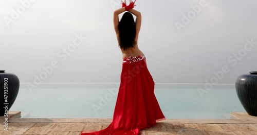 A beautiful belly dancer in a red costume performing front of the pool and open sea, smoothly moving showing curves of the waist in motion - Back view  photo