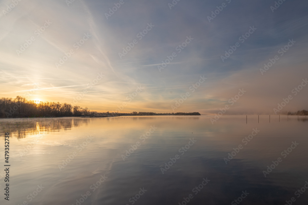 Early winter morning at a lake during a sunrise and the sunlight reflects over the water of the lake and creates a fairy tale landscape together with the frozen ground and grass and a veil of mist