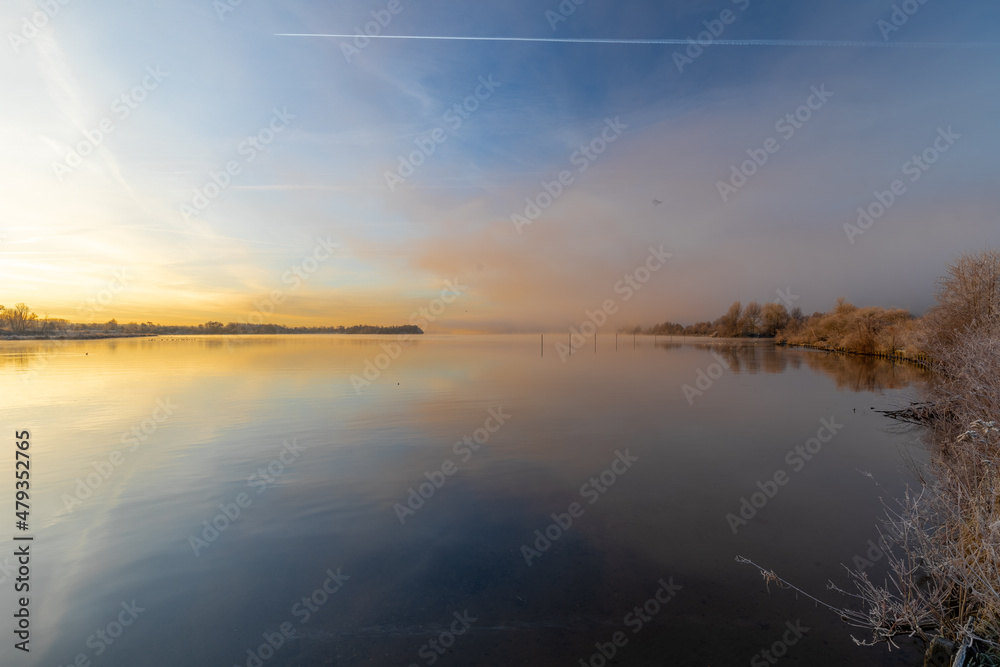 Early winter morning at a lake during a sunrise and the sunlight reflects over the water of the lake and creates a fairy tale landscape together with the frozen ground and grass and a veil of mist