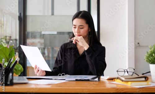 Young asian business woman sitting at her workplace and reading financial document.