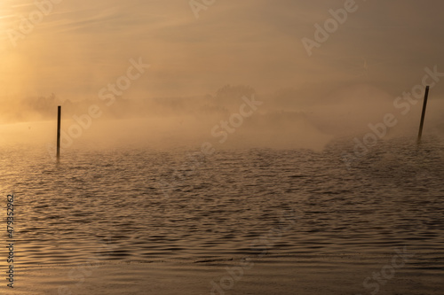 Early winter morning at a lake during a sunrise and the sunlight reflects over the water of the lake and creates a fairy tale landscape together with the frozen ground and grass and a veil of mist