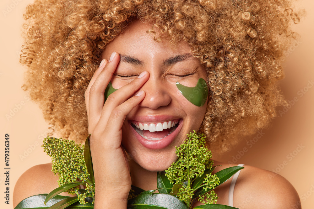 Beauty and skin care concept. Overjoyed woman laughs out happily covers face with hand smiles toothily has natural clean skin applies green collagen patches under eyes for reducing wrinkles. - obrazy, fototapety, plakaty 