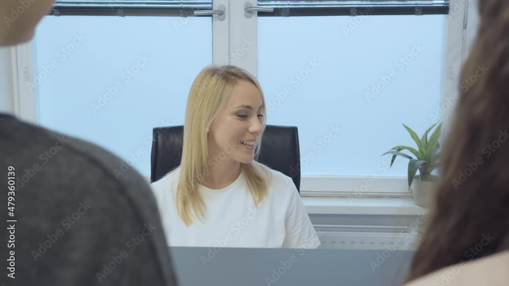 Over the shoulder of two female patients standing at the reception desk in a medical office and talking with a Caucasian female receptionist.