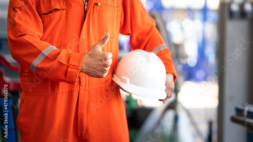 Obraz na plátne A worker in orange coverall is thumb up action and holding white safety helmet with factory workshop as blurred background