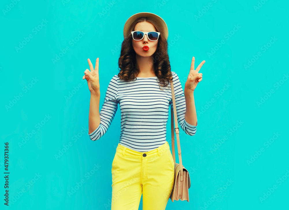 Portrait of beautiful young woman blowing her lips sending sweet air kiss posing wearing a handbag, striped t-shirt and summer straw round hat on blue background