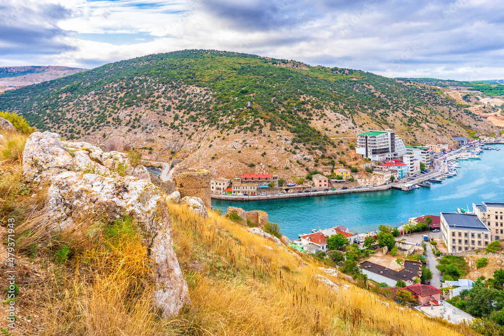 Panoramic view from hill to sea bay and small coastal town in mountain area. Picturesque urban landscape in summer cloudy day. Balaklava in Sevastopol vicinity, Crimea, Russia