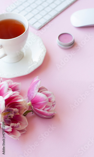 Three pink peony tulips and cup of black tea, white wireless keyboard and mouse, lips balm on pink background