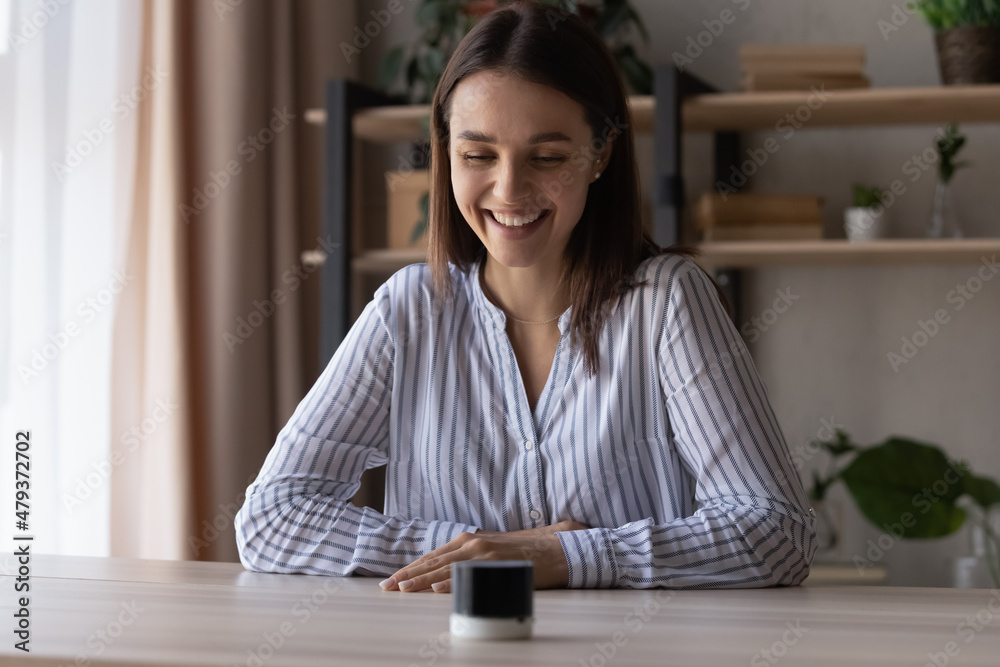 Happy young woman using small portable speaker, looking at gadget on table, smiling, laughing. Millennial customer giving command, talking to voice assistant. Modern smart home technology Happy young woman using small portable speaker, looking at gadget on table, smiling, laughing. Millennial customer giving command, talking to voice assistant. Modern smart home technology