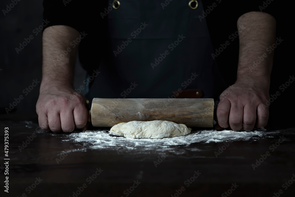 Man Baker or chef is rolling fresh dough with a dough roller on a wooden surface.