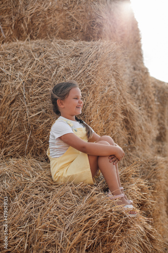 Portrait of sitting girl on high haystack smiling with teeth looking somewhere far away wearing sundress. Having fun away from city on field full of golden hay.