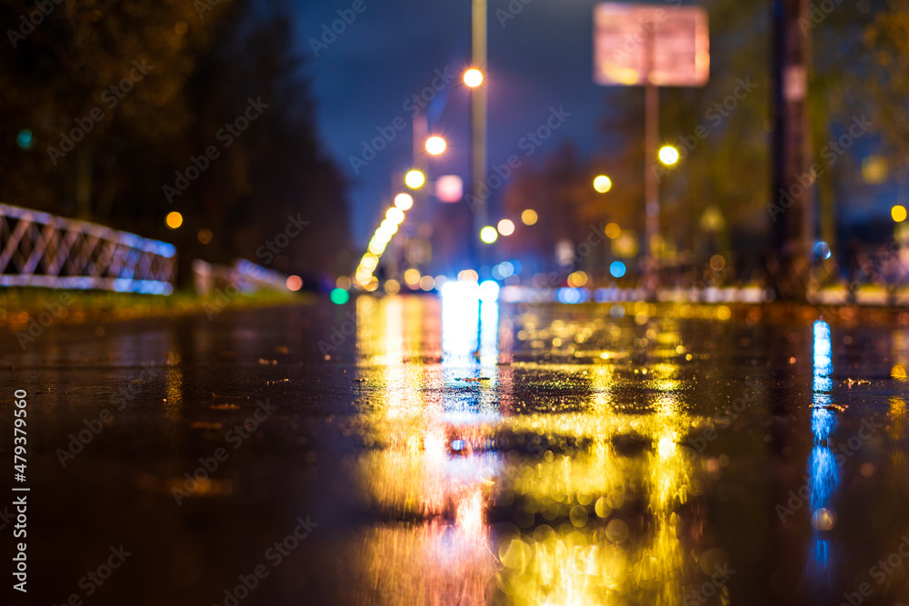 Night city in the rain. The light from the lanterns reflects on the wet pedestrian sidewalk. Falling drops of rain. Colorful colors. Focus on the asphalt. Close up view from the asphalt level.