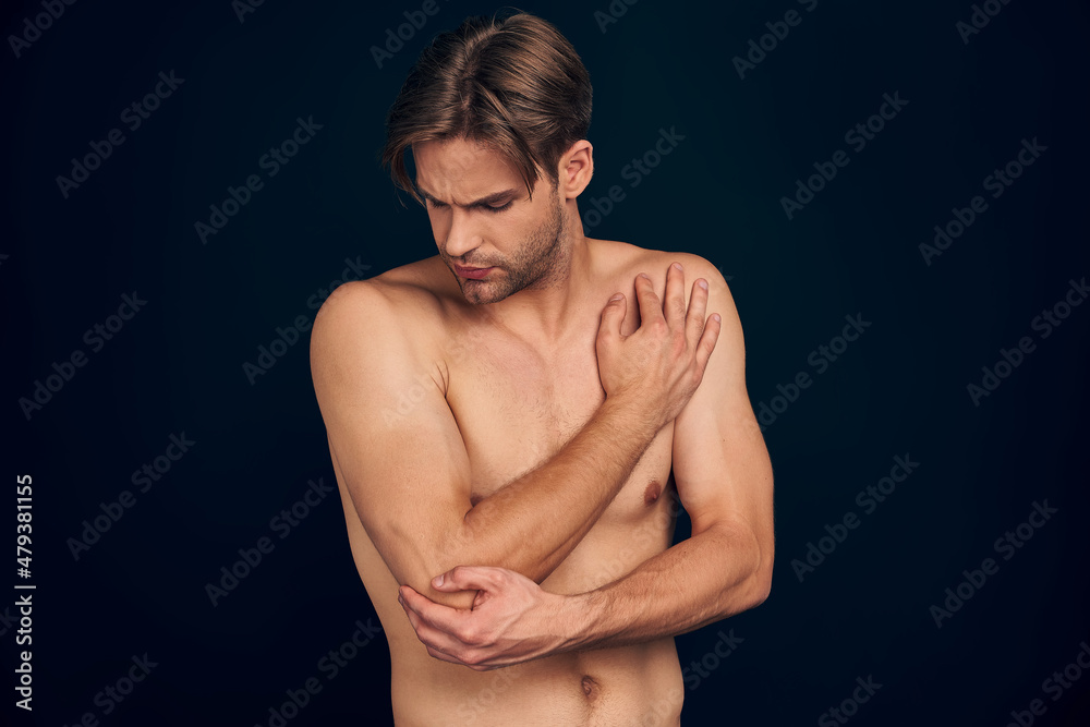 Handsome young bearded man isolated. Cropped image of shirtless muscular man is standing on dark blue background. Man holding his elbow. Experiencing elbow pain