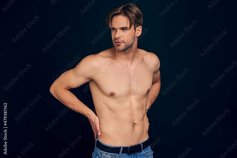Handsome young bearded man isolated. Portrait of shirtless muscular man is standing on grey background and holding his back