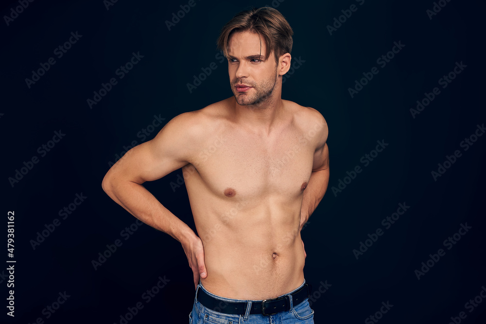 Handsome young bearded man isolated. Portrait of shirtless muscular man is standing on grey background and holding his back Handsome young bearded man isolated. Portrait of shirtless muscular man is standing on grey background and holding his back