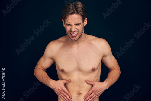Handsome young bearded man isolated. Cropped image of shirtless muscular man is standing on dark blue background. Man holding his stomach with big scars. Experiencing stomach pain